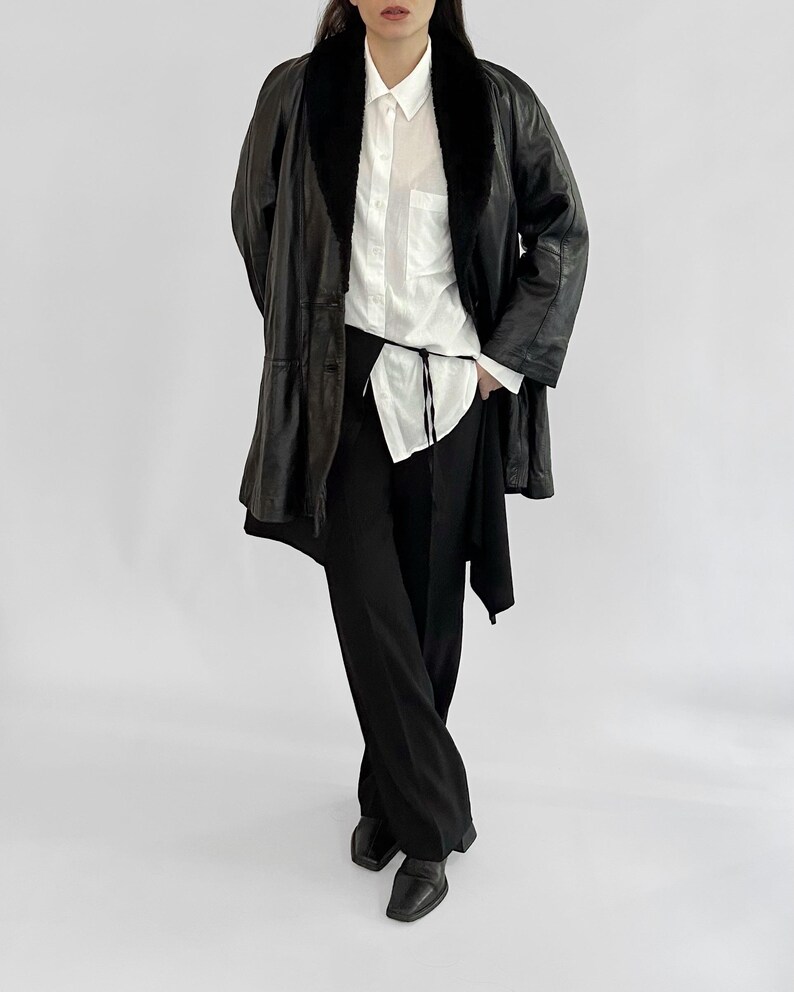 May include: A black leather coat with a faux fur collar, worn over a white button-down shirt and black pants. The coat has a long, flowing design. The outfit is completed with black shoes.