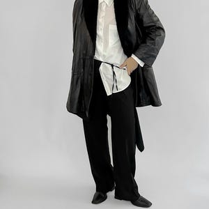 May include: A black leather coat with a faux fur collar, worn over a white button-down shirt and black pants. The coat has a belted waist and a classic, tailored design. The outfit is completed with black shoes.