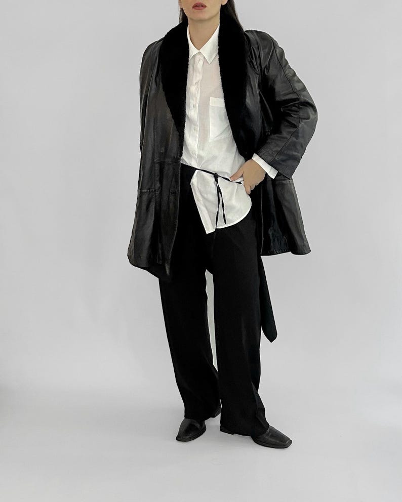 May include: A black leather coat with a faux fur collar, worn over a white button-down shirt and black pants. The coat has a belted waist and a classic, tailored design. The outfit is completed with black shoes.