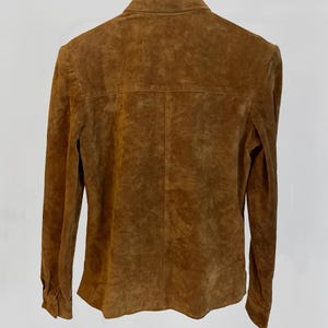 May include: A brown suede jacket with long sleeves and a buttoned front. The jacket is hanging on a black hanger against a white background. The suede has a textured appearance.