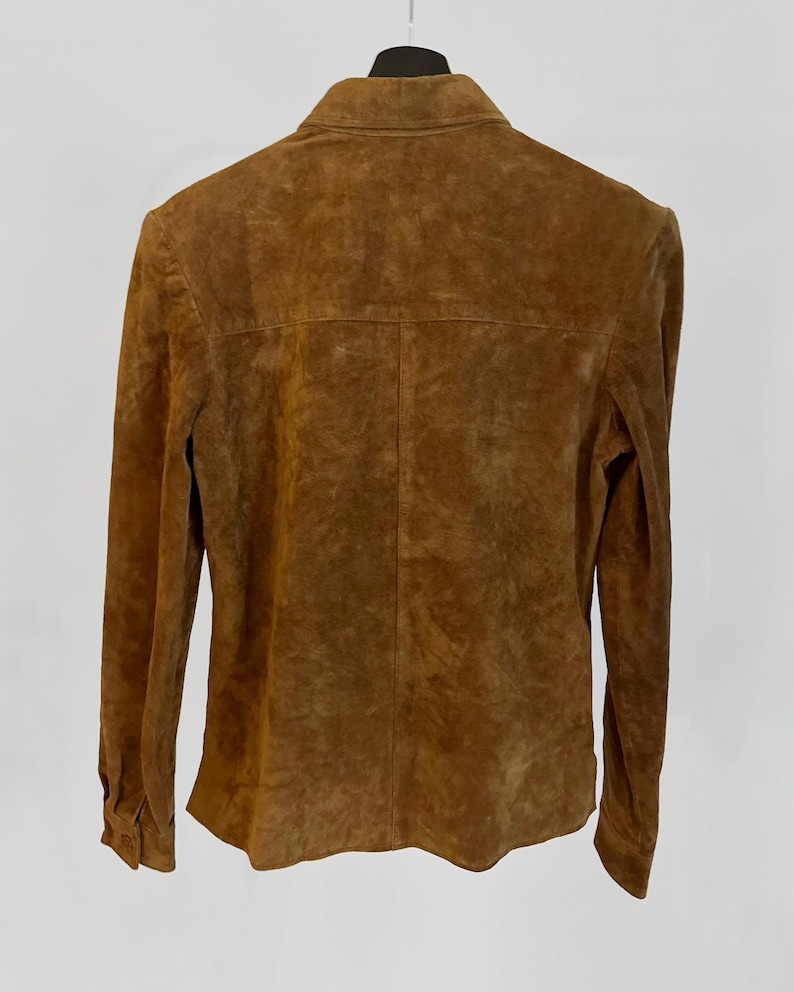 May include: A brown suede jacket with long sleeves and a buttoned front. The jacket is hanging on a black hanger against a white background. The suede has a textured appearance.