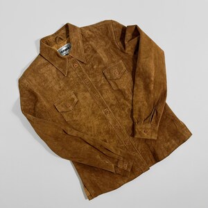 May include: A brown suede shirt with two chest pockets and button closures. The shirt has a collared neckline and long sleeves with buttoned cuffs. The garment is laid flat on a white surface.