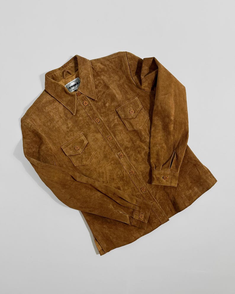 May include: A brown suede shirt with two chest pockets and button closures. The shirt has a collared neckline and long sleeves with buttoned cuffs. The garment is laid flat on a white surface.