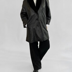 May include: A black leather coat with a black fur collar and three buttons. The coat is worn over a white collared shirt and black pants. The outfit is completed with black shoes.