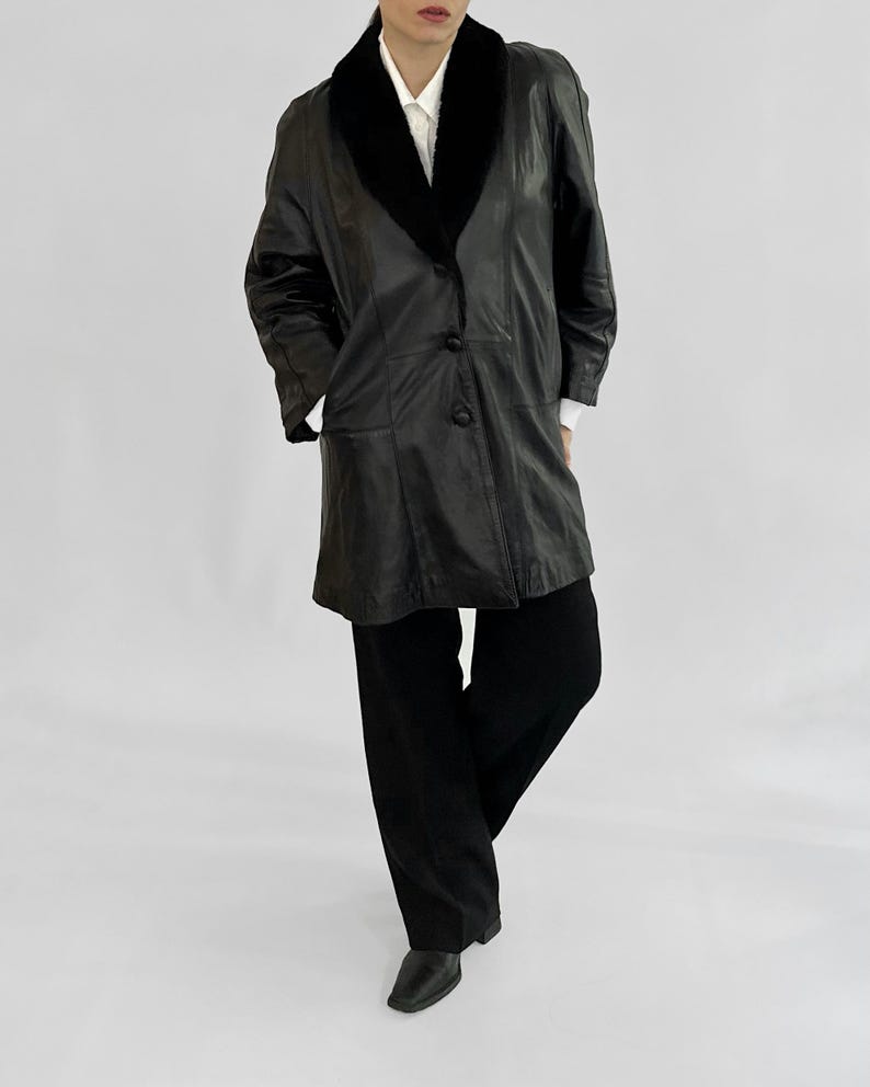 May include: A black leather coat with a black fur collar and three buttons. The coat is worn over a white collared shirt and black pants. The outfit is completed with black shoes.