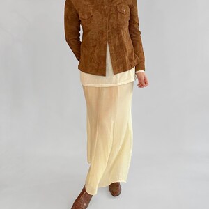 May include: A brown suede button-up shirt with two chest pockets, paired with a cream-colored top and a long, flowing, pale yellow skirt. The outfit is completed with brown leather boots.