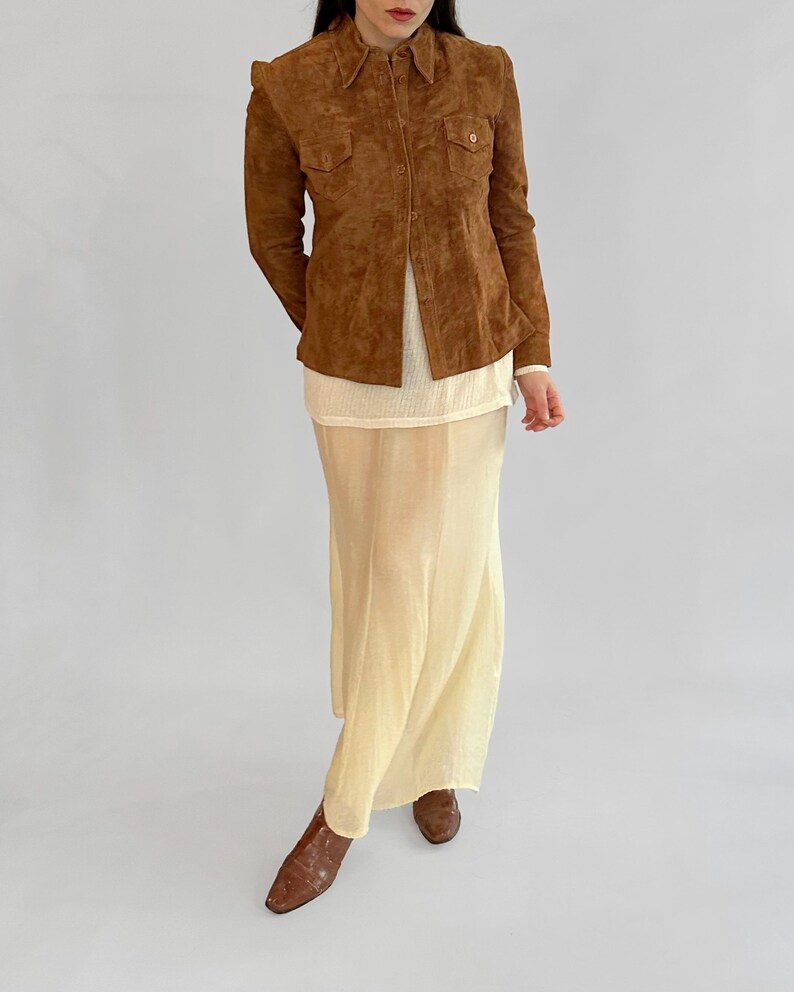 May include: A brown suede button-up shirt with two chest pockets, paired with a cream-colored top and a long, flowing, pale yellow skirt. The outfit is completed with brown leather boots.