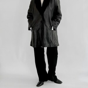 May include: A black leather coat with a black faux fur collar. The coat has a button closure and two front pockets. The person is wearing black pants and black shoes. The background is a plain white.
