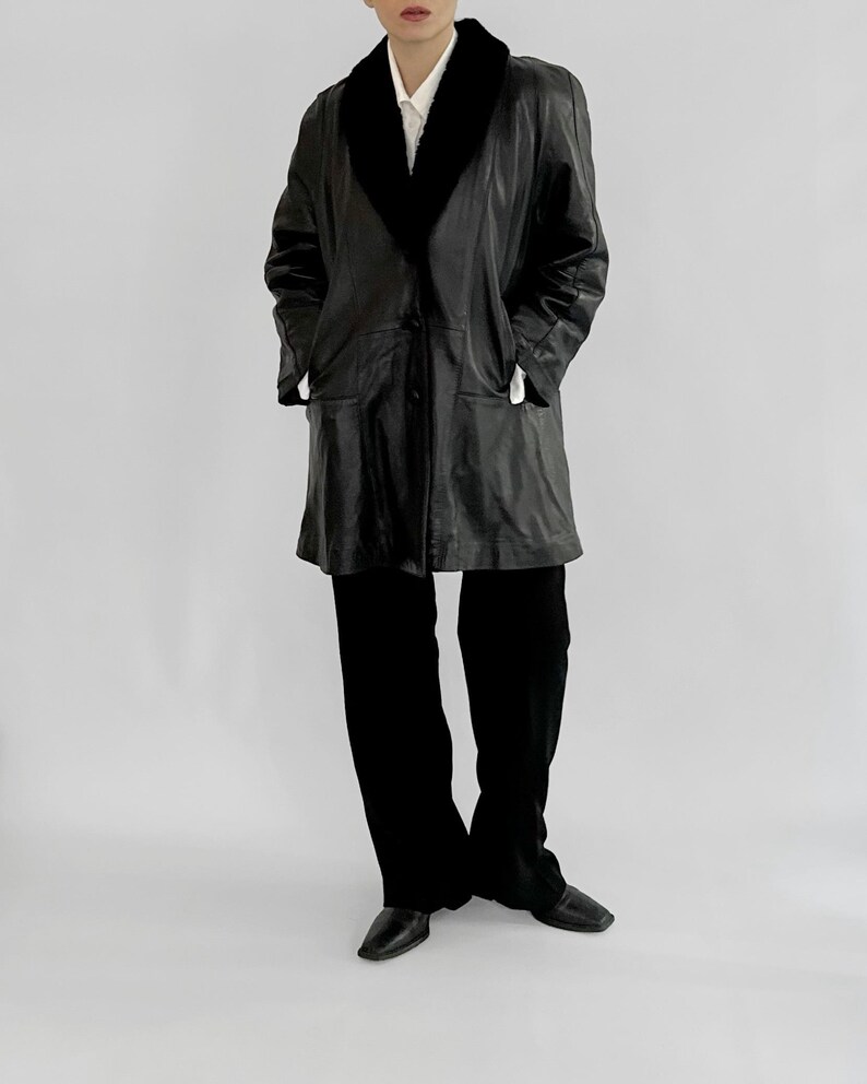 May include: A black leather coat with a black faux fur collar. The coat has a button closure and two front pockets. The person is wearing black pants and black shoes. The background is a plain white.