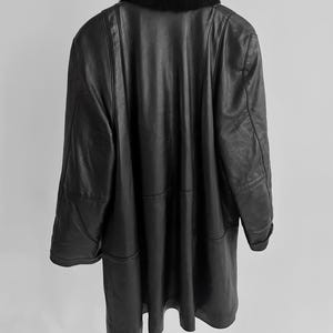 May include: A long, black leather coat with a black fur collar hangs from a hanger. The coat has a classic design with a tailored fit and visible seams. The coat is a vintage item.