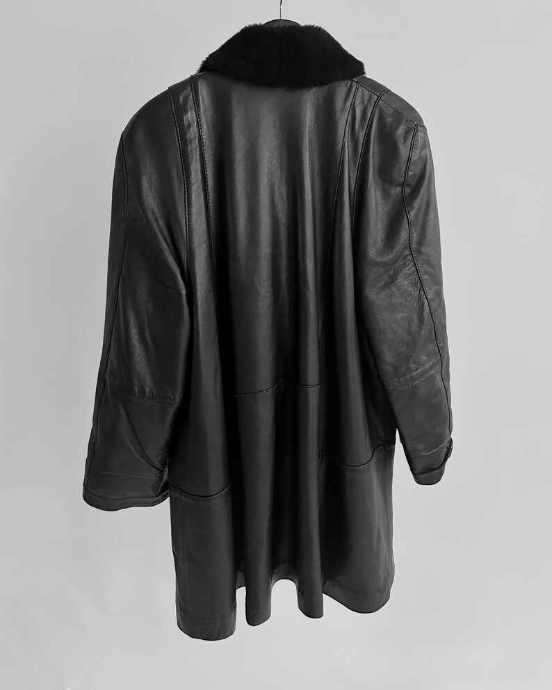 May include: A long, black leather coat with a black fur collar hangs from a hanger. The coat has a classic design with a tailored fit and visible seams. The coat is a vintage item.