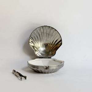 May include: A silver-toned metal clam shell-shaped container with a hinged lid that opens to reveal a white interior. The container features a ribbed exterior texture and is accompanied by a small, matching silver-toned metal pair of tweezers.