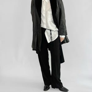 May include: A black leather jacket with a faux fur collar, worn over a white button-down shirt and black pants. The jacket has a long, tailored silhouette. The outfit is completed with black shoes.