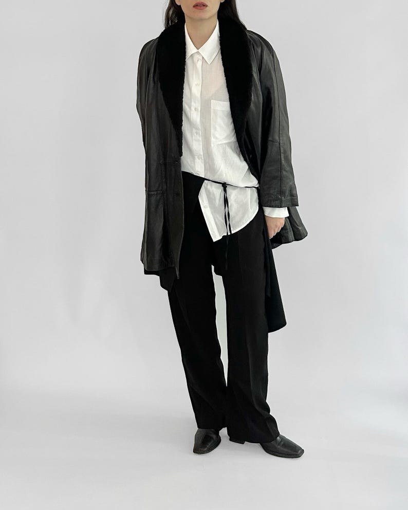 May include: A black leather jacket with a faux fur collar, worn over a white button-down shirt and black pants. The jacket has a long, tailored silhouette. The outfit is completed with black shoes.