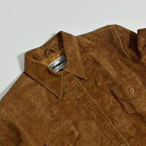 May include: A brown suede shirt with a collared neckline, button-down front, and two chest pockets. The shirt has a vintage look and feel. The brand tag is visible at the collar.