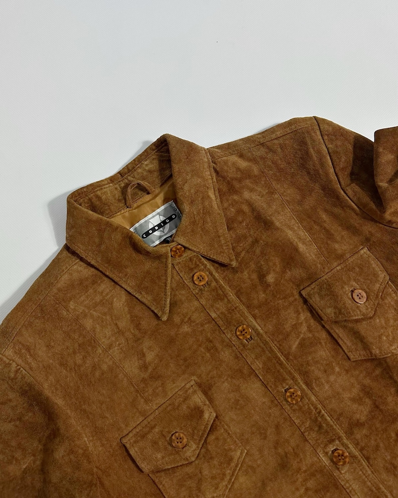 May include: A brown suede shirt with a collared neckline, button-down front, and two chest pockets. The shirt has a vintage look and feel. The brand tag is visible at the collar.