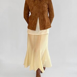 May include: A brown suede jacket with a collared neckline and two chest pockets. The jacket is paired with a cream-colored top and a long, flowing, pale yellow skirt. Brown boots complete the outfit.