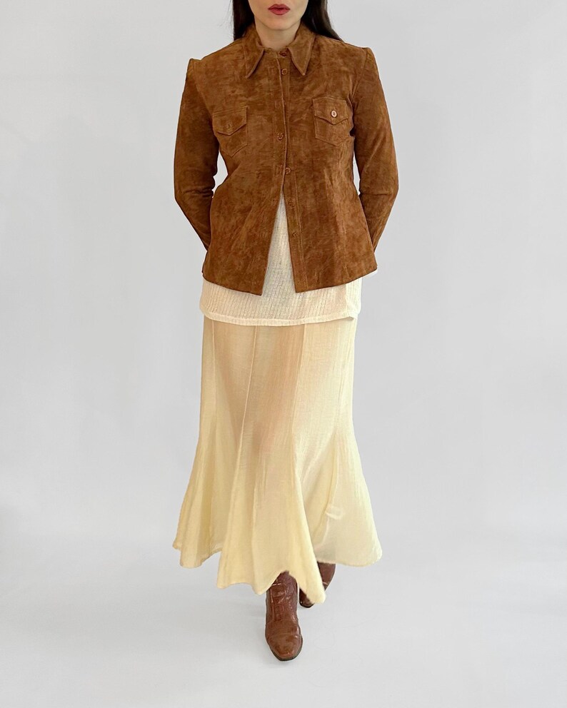 May include: A brown suede jacket with a collared neckline and two chest pockets. The jacket is paired with a cream-colored top and a long, flowing, pale yellow skirt. Brown boots complete the outfit.