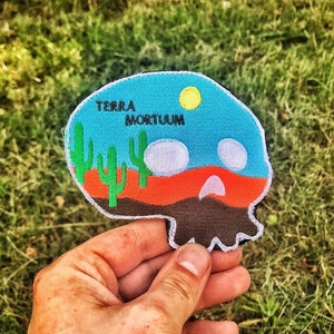 Op de afbeelding: Een blauwe en oranje geborduurde patch met een schedelontwerp. De schedel is gevuld met een woestijnlandschap met cactussen en een zon. De tekst "TERRA MORTUUM" staat op de patch.