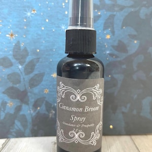 Könnte beinhalten: Dunkle Glassprühflasche mit schwarzem Verschluss und transparentem Kunststoffaufsatz. Das Etikett lautet "Cinnamon Broom Spray" mit "Abundance & Prosperity" darunter. Die Flasche enthält 59 ml Flüssigkeit.