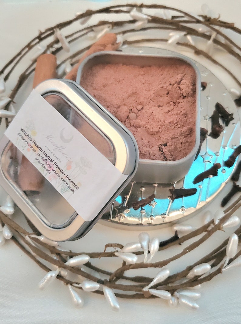 May include: A silver tin with a clear lid containing a brown herbal powder incense blend. The label reads "Witches Hearth Herbal Powder Incense" with a list of ingredients including cinnamon, love, sandalwood, arjuna, clove buds, and essential oils.