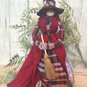 Puede incluir: Muñeca de bruja hecha a mano con sombrero negro y vestido a cuadros rojos. La muñeca tiene una cabeza de madera y sostiene una pequeña escoba. El atuendo de la bruja incluye una larga capa roja y un cinturón negro. Es un artículo decorativo.