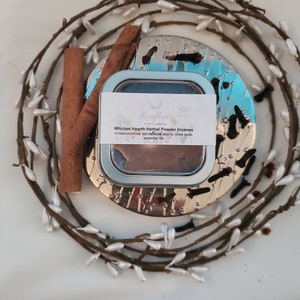May include: A round tin container with a silver lid and a white label that reads "Witches Hearth Herbal Powder Incense" with a list of ingredients: cinnamon, clove, sandalwood, arjuna, clove buds, essential oils. The tin is sitting on a silver tray with a wreath of white berries around it.
