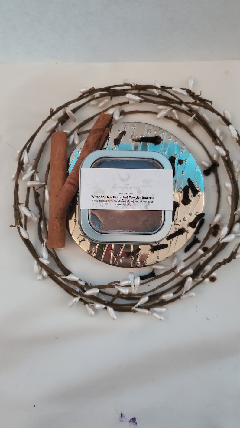 May include: A round tin container with a silver lid and a white label that reads "Witches Hearth Herbal Powder Incense" with a list of ingredients: cinnamon, clove, sandalwood, arjuna, clove buds, essential oils. The tin is sitting on a silver tray with a wreath of white berries around it.