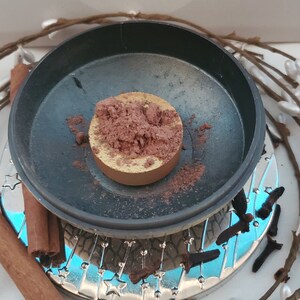 May include: A round, gold-colored metal container filled with a brown powder. The container is sitting on a black surface with a silver rim. The container is surrounded by cinnamon sticks and cloves.