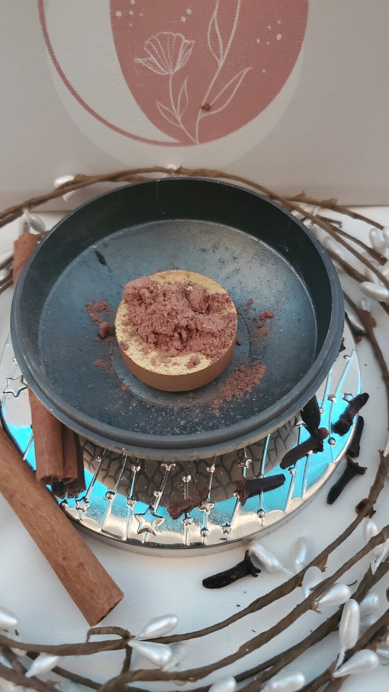 May include: A round, gold-colored metal container filled with a brown powder. The container is sitting on a black surface with a silver rim. The container is surrounded by cinnamon sticks and cloves.