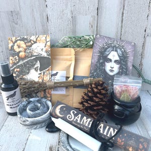 May include: A collection of items including a black spray bottle labeled Harvest Moon, a gray marbled candle holder, a pinecone, a small black cauldron, a white candle, a silver dish, and two cards featuring illustrations of a woman and a figure.