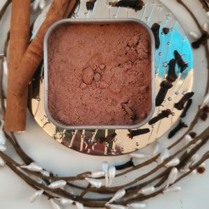 May include: A small metal tin filled with a brown powder, surrounded by cinnamon sticks and cloves. The tin is sitting on a silver tray with a decorative design.