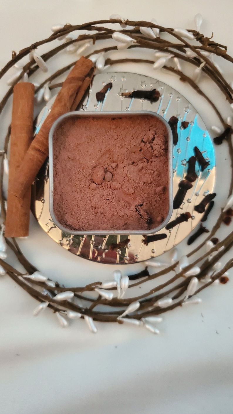 May include: A small metal tin filled with a brown powder, surrounded by cinnamon sticks and cloves. The tin is sitting on a silver tray with a decorative design.
