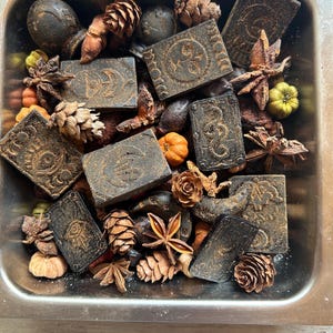 以下が含まれることがあります： 金属製の容器に、松ぼっくり、八角、小さなカボチャに囲まれた、エンボス加工が施された濃い茶色の長方形の石鹸が入っています。石鹸には様々なシンボルや模様が施されています。