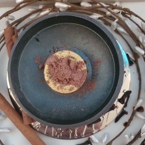 May include: A black metal incense burner with a gold rim and a pile of brown incense powder inside. The burner is surrounded by a wreath of white berries and brown twigs.
