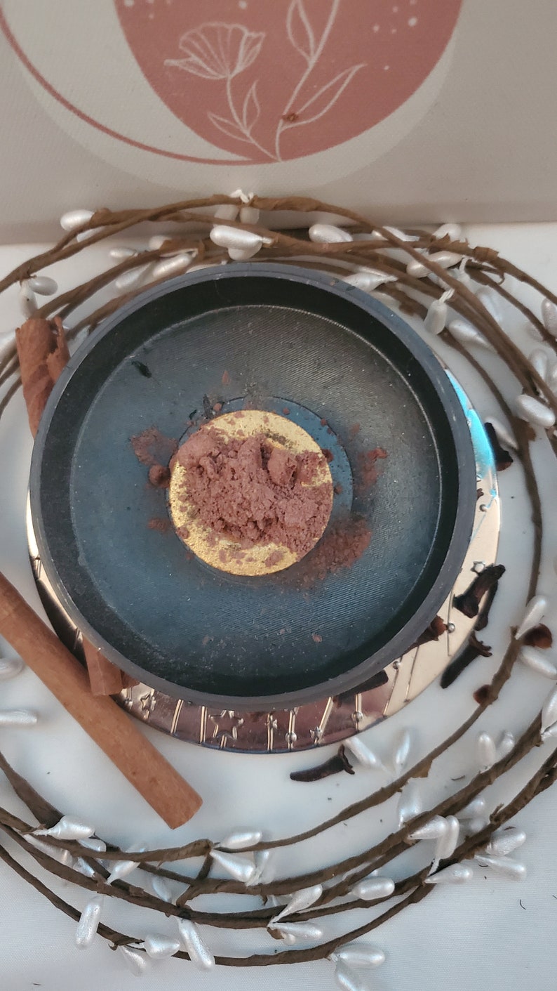 May include: A black metal incense burner with a gold rim and a pile of brown incense powder inside. The burner is surrounded by a wreath of white berries and brown twigs.
