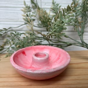 Op de afbeelding: Een roze hars kaarsenhouder met een marmer effect. De houder heeft een cirkelvormige basis met een verhoogd midden voor een kaars. De achtergrond omvat groen en een houten oppervlak.