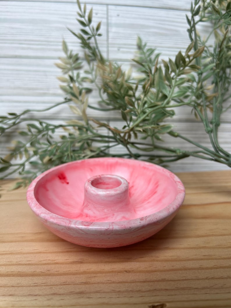Op de afbeelding: Een roze hars kaarsenhouder met een marmer effect. De houder heeft een cirkelvormige basis met een verhoogd midden voor een kaars. De achtergrond omvat groen en een houten oppervlak.