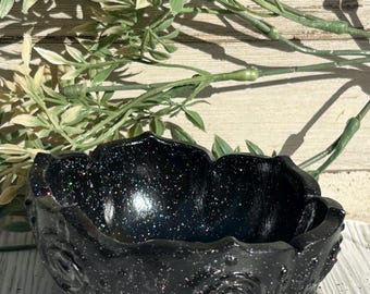 Puede incluir: Un cuenco de resina negra con purpurina y diseño floral. El cuenco tiene una superficie texturizada y tiene forma de flor. El cuenco está sobre una superficie de madera con follaje verde en el fondo.