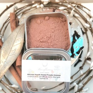 May include: A small tin container filled with a brown herbal powder incense blend. The label reads "Moonflower's Mystic Garden Witches Hearth Herbal Powder Incense" and lists the ingredients: cinnamon, clove, sandalwood, arjuna, clove buds, and essential oils.