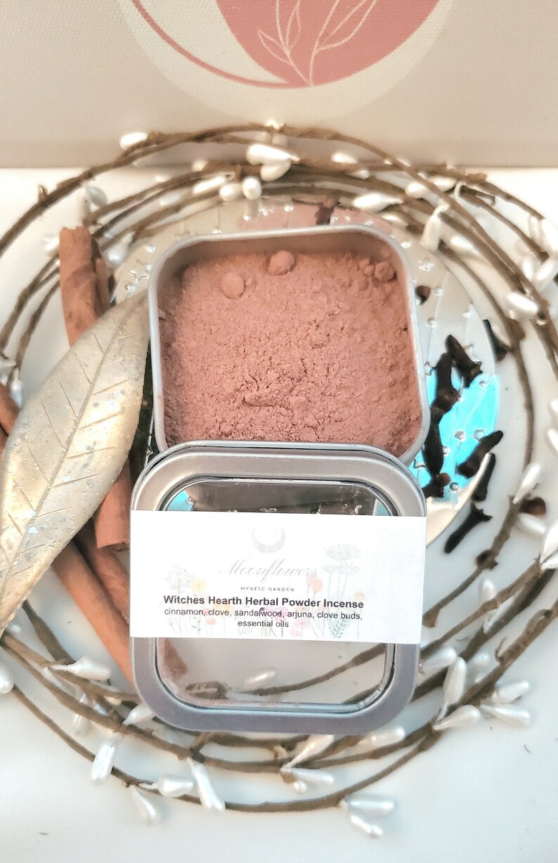 May include: A small tin container filled with a brown herbal powder incense blend. The label reads "Moonflower's Mystic Garden Witches Hearth Herbal Powder Incense" and lists the ingredients: cinnamon, clove, sandalwood, arjuna, clove buds, and essential oils.