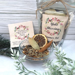 May include: A clear plastic bag filled with a simmer pot blend for the Yule season. The bag is tied with twine and has a label that reads "Yule Blessings Simmer Pot". The label features a watercolor illustration of a wreath with red berries and green leaves. A small bowl filled with the simmer pot blend is next to the bag. The ingredients include orange peel, cinnamon sticks, cloves, bay leaves, and allspice.