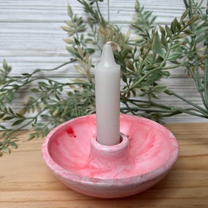Op de afbeelding: Een roze en witte gemarmerde kandelaar met een witte kaars. De kandelaar is rond met een centrale opening voor de kaars. De kaars is wit en niet aangestoken. De achtergrond omvat groen en een houten oppervlak.