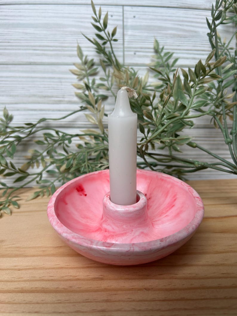 Op de afbeelding: Een roze en witte gemarmerde kandelaar met een witte kaars. De kandelaar is rond met een centrale opening voor de kaars. De kaars is wit en niet aangestoken. De achtergrond omvat groen en een houten oppervlak.