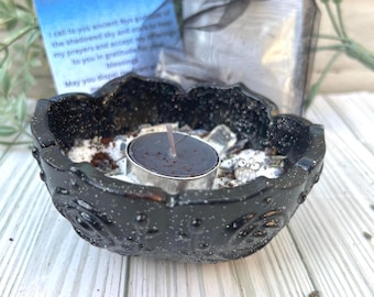 May include: A black speckled decorative bowl with a lit black candle, surrounded by white and brown granules. A small card with text and a black organza bag are in the background. The bowl has a floral design.