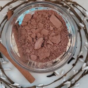May include: A close-up of a small glass jar filled with a brown powder. The powder is a light brown color and has a slightly grainy texture. The jar is sitting on a white surface.