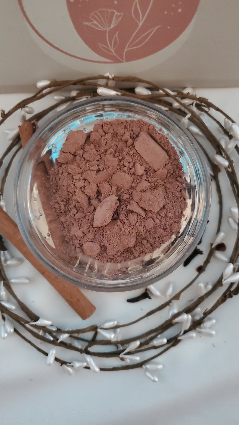 May include: A close-up of a small glass jar filled with a brown powder. The powder is a light brown color and has a slightly grainy texture. The jar is sitting on a white surface.