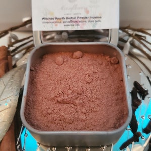 May include: A silver tin with a brown powder inside. The label on the tin reads "Witches Hearth Herbal Powder Incense" and lists the ingredients: cinnamon, clove, sandalwood, arjuna, clove buds, and essential oils.
