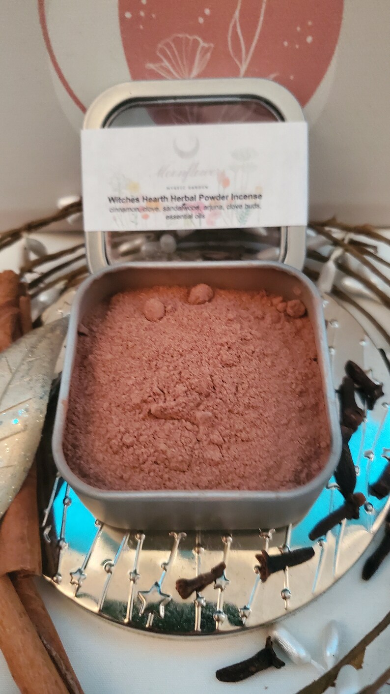 May include: A silver tin with a brown powder inside. The label on the tin reads "Witches Hearth Herbal Powder Incense" and lists the ingredients: cinnamon, clove, sandalwood, arjuna, clove buds, and essential oils.