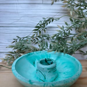 Op de afbeelding: Een teal en wit gemarmerde betonnen kandelaar. De kandelaar heeft een cirkelvormige basis met een verhoogd midden voor de kaars. De achtergrond omvat groen en een wit houten oppervlak.